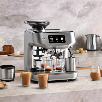 Silver Breville coffee machine on a kitchen counter with glasses of coffee and a kettle.