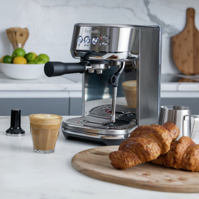 Stainless steel espresso machine with Breville branding, angled profile with croissants, cortado in a glass cup, and tamper in front
