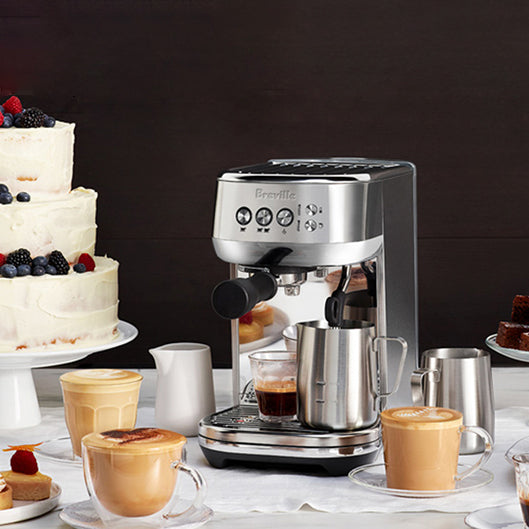 Stainless steel espresso machine with Breville branding, angled profile surrounded by pastries and lattes in glass cups