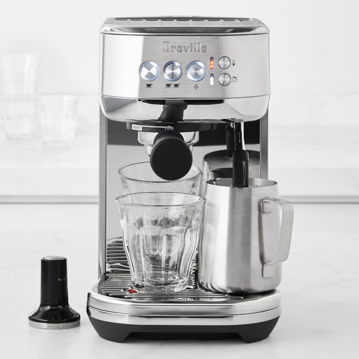 Stainless steel espresso machine with Breville branding with a glass cup, frothing pitcher, and tamper in front
