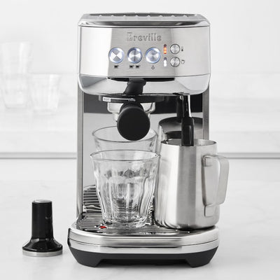 Stainless steel espresso machine with Breville branding with a glass cup, frothing pitcher, and tamper in front