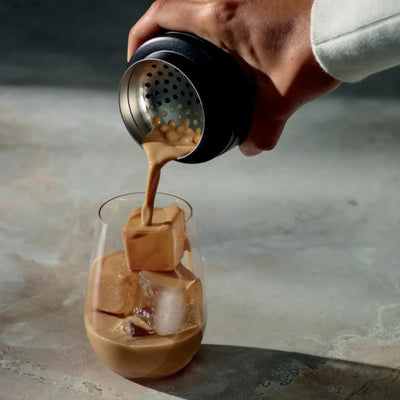 Hand pouring a coffee from a Breville The Shake & Take Black Truffle - BEA050BTR0ZNA1 into a glass with ice cubes on a marble surface.
