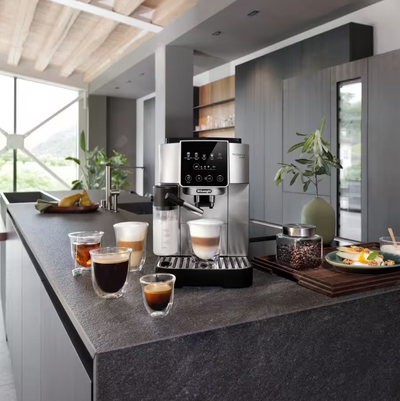A modern kitchen countertop setup featuring a stainless steel and black coffee machine dispensing a latte, with several small cups of espresso and coffee drinks arranged nearby. The scene is set in a contemporary kitchen with dark gray cabinets, a wood ceiling, and large windows.