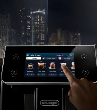 Person interacting with a Delonghi coffee machine's touchscreen interface at night with city skyline in the background.