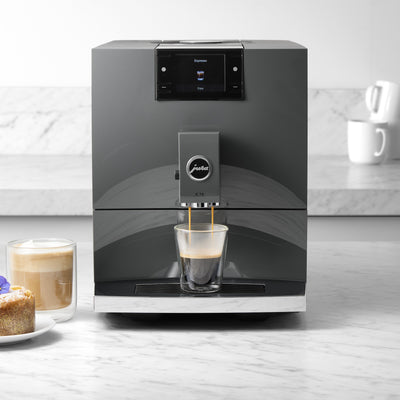 Modern coffee machine dispensing a shot of espresso next to a glass of coffee and a cake on a kitchen counter.