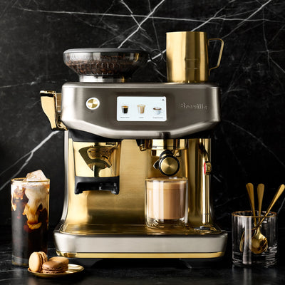 Gold and silver Breville espresso machine on a dark marble background with coffee accessories.