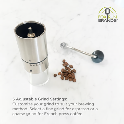 A stainless steel manual coffee grinder with a clear lower chamber and numbered grind settings, shown on a white marble surface with coffee beans and a metal lever. Text explains the 5 adjustable grind settings, and the Fox Run Brands logo appears in the top right.