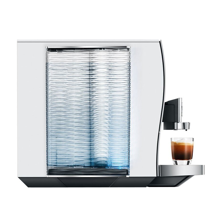 A sleek white and stainless steel coffee machine with a textured glass water tank, shown with a glass of espresso on its drip tray against a solid white background.