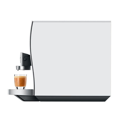 A modern coffee machine with a large white front panel, black base, and a glass of espresso with milk on the drip tray, shown against a solid white background.