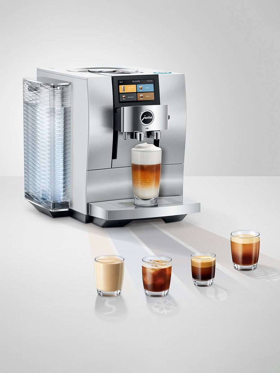A stainless steel coffee machine with a digital touch screen and a water tank on the left, shown dispensing a layered latte into a glass. Four small glasses of coffee or milk-based drinks are lined up in front on a light gray reflective surface against a neutral studio background.