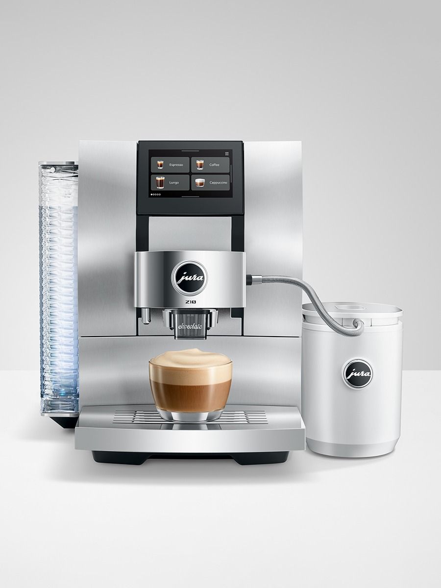 A sleek jura automatic espresso machine in stainless steel, shown with a glass cup of cappuccino on its drip tray, a tall water reservoir on the left, and a white milk container on the right, set against a neutral light gray background.