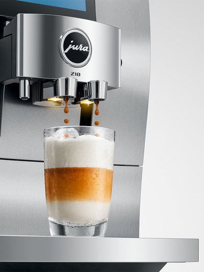 A Jura Z10 espresso machine dispensing coffee into a glass of layered latte with milk foam, shown against a clean metallic and light gray background.