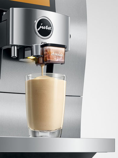 A close-up of a Jura Z10 coffee machine dispensing espresso into a clear glass cup filled with milk, creating a creamy latte. The machine features a brushed stainless steel body with the Jura logo clearly visible.