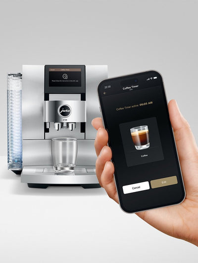 A sleek Jauslé coffee machine in stainless steel shown next to a hand holding a smartphone displaying a coffee timer app on a light gray background.